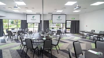 Un salón de conferencias con varias mesas y sillas, ventanas amplias con cortinas y una pantalla que muestra el logotipo de Holiday Inn.
