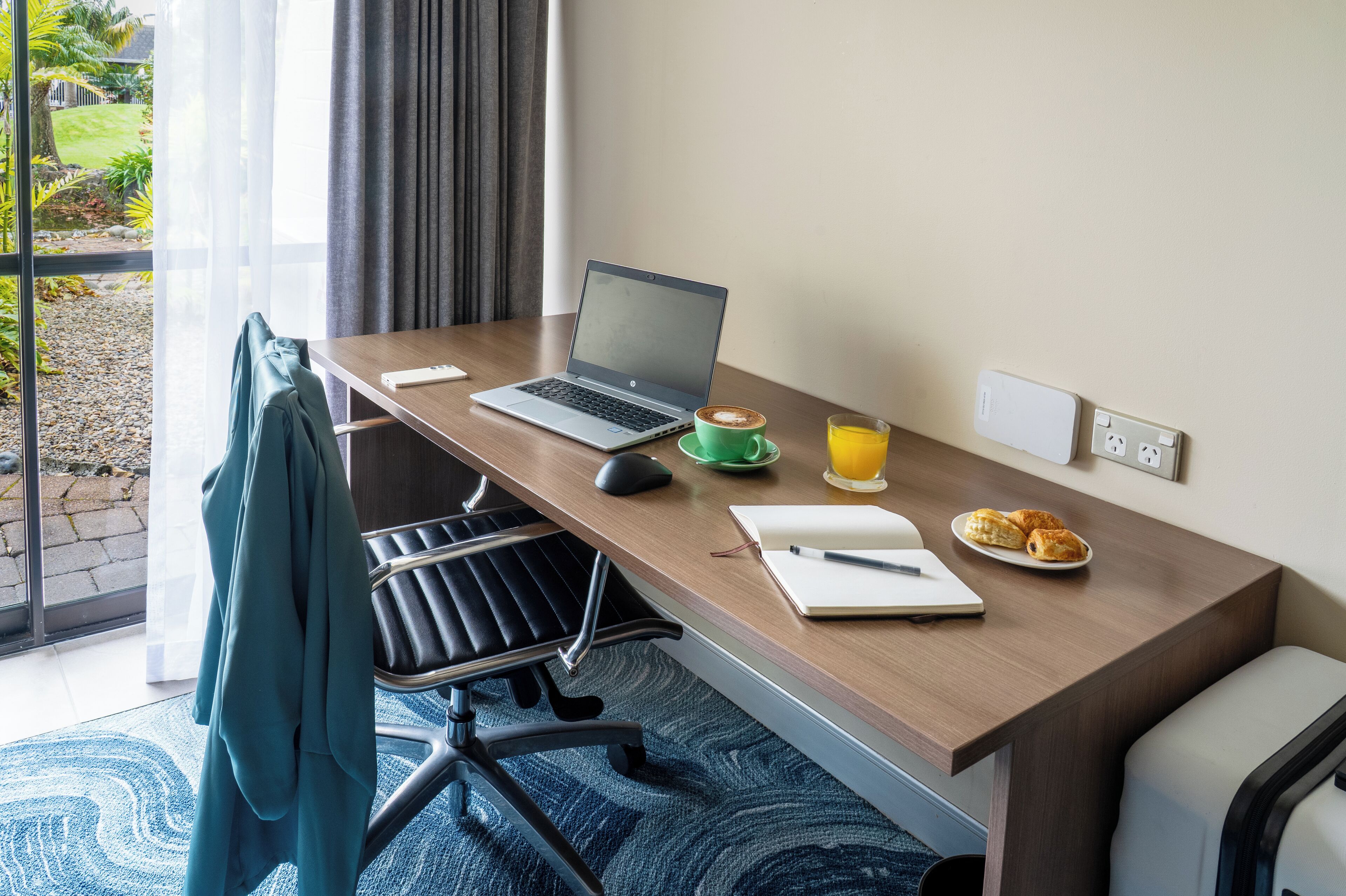 Habitación de hotel con escritorio de madera, computadora portátil, libreta, bolígrafo, taza verde, jugo de naranja y croissants.