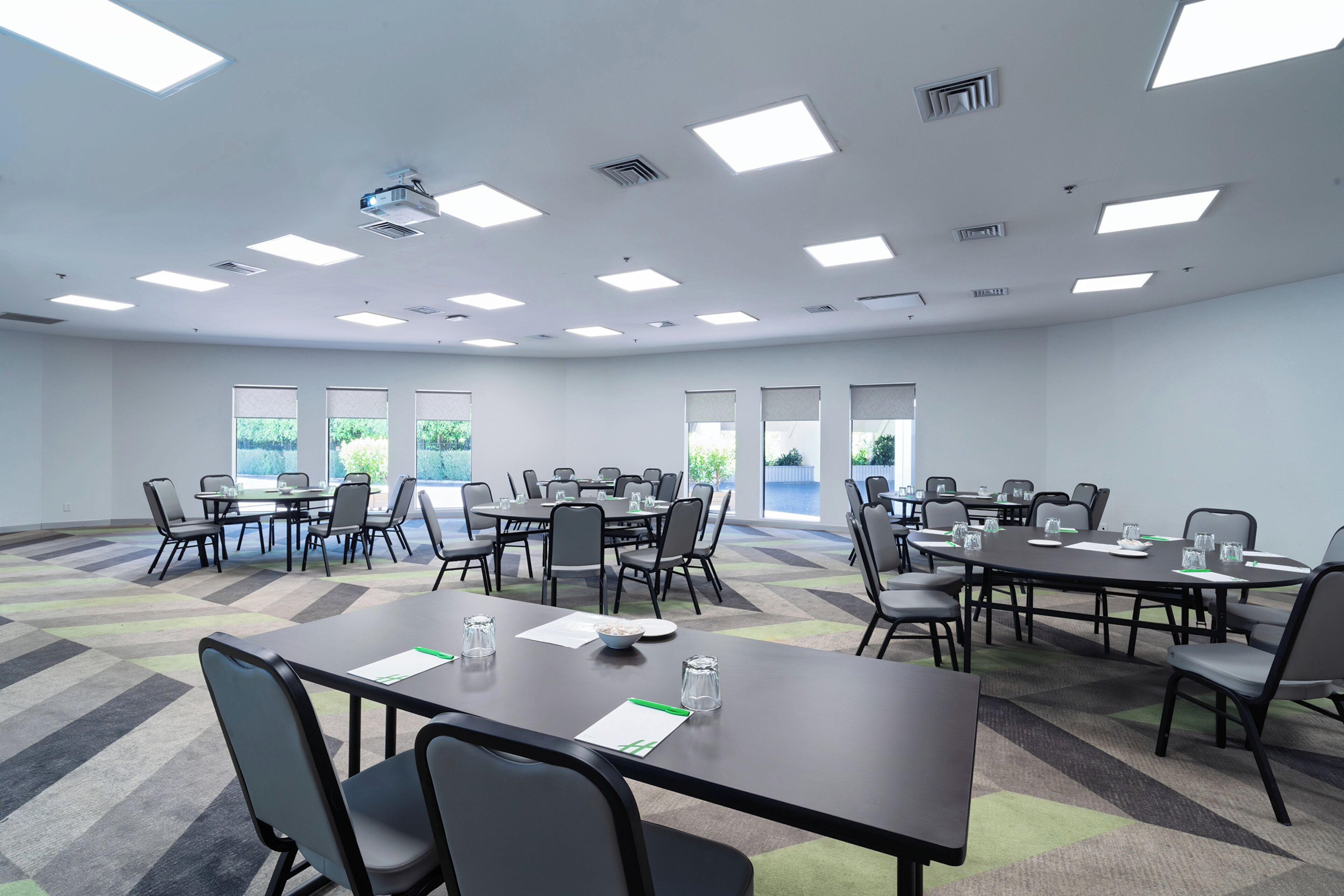 Un salón de conferencias con varias mesas y sillas, ventanas amplias e iluminación en el techo.