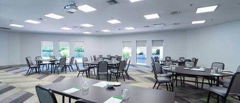Un salón de conferencias con varias mesas y sillas, ventanas amplias e iluminación en el techo.