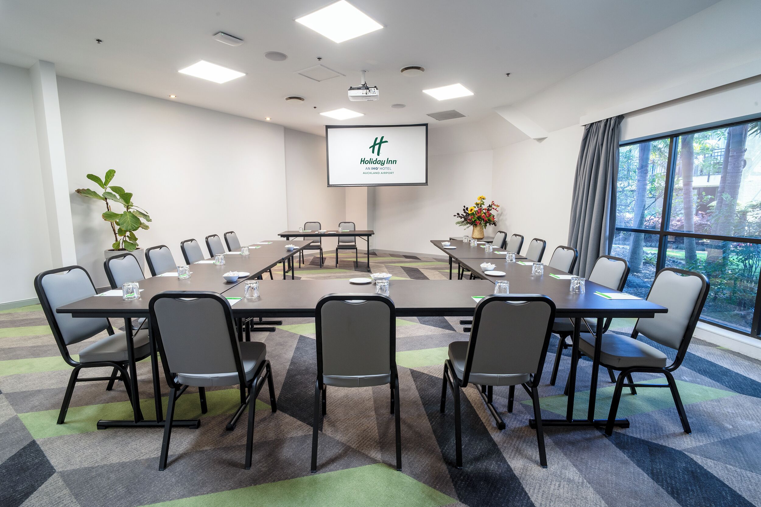 A conference room with a large table, chairs, a projector screen, and a view of trees through a window.