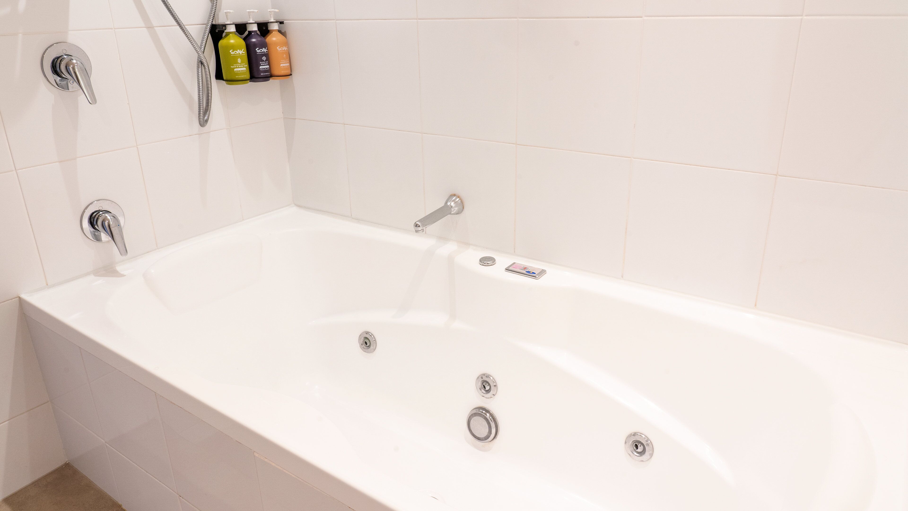 A white bathtub with a handheld showerhead, a built-in faucet, and a handheld shower attachment. There are three bottles of soap on a wall-mounted soap dispenser.