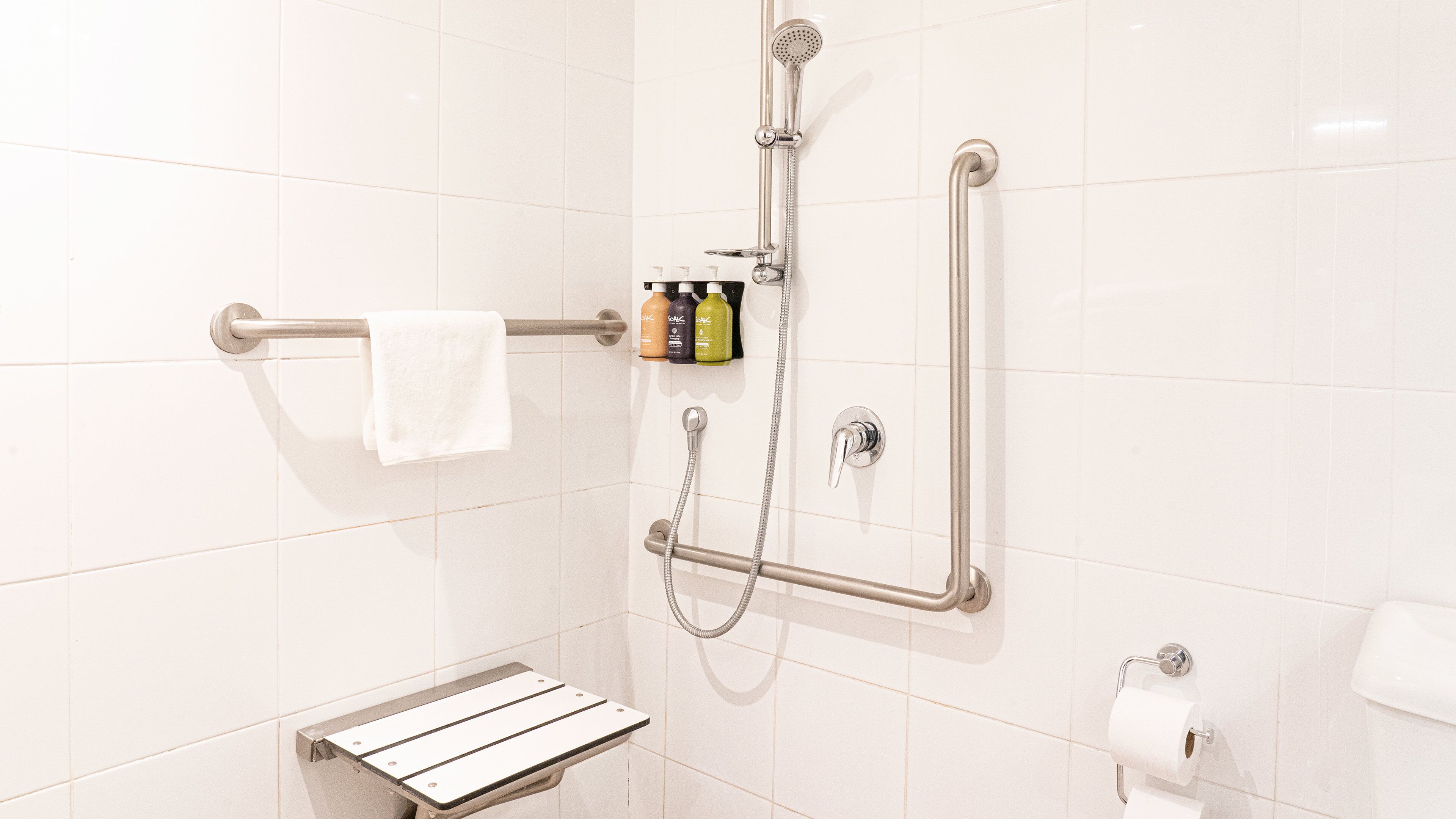 Une salle de bain avec des murs recouverts de carreaux blancs, une douche équipée d’un pommeau de douche à main, d’une barre d’appui, d’un porte-serviettes et d’une étagère contenant des articles de toilette.