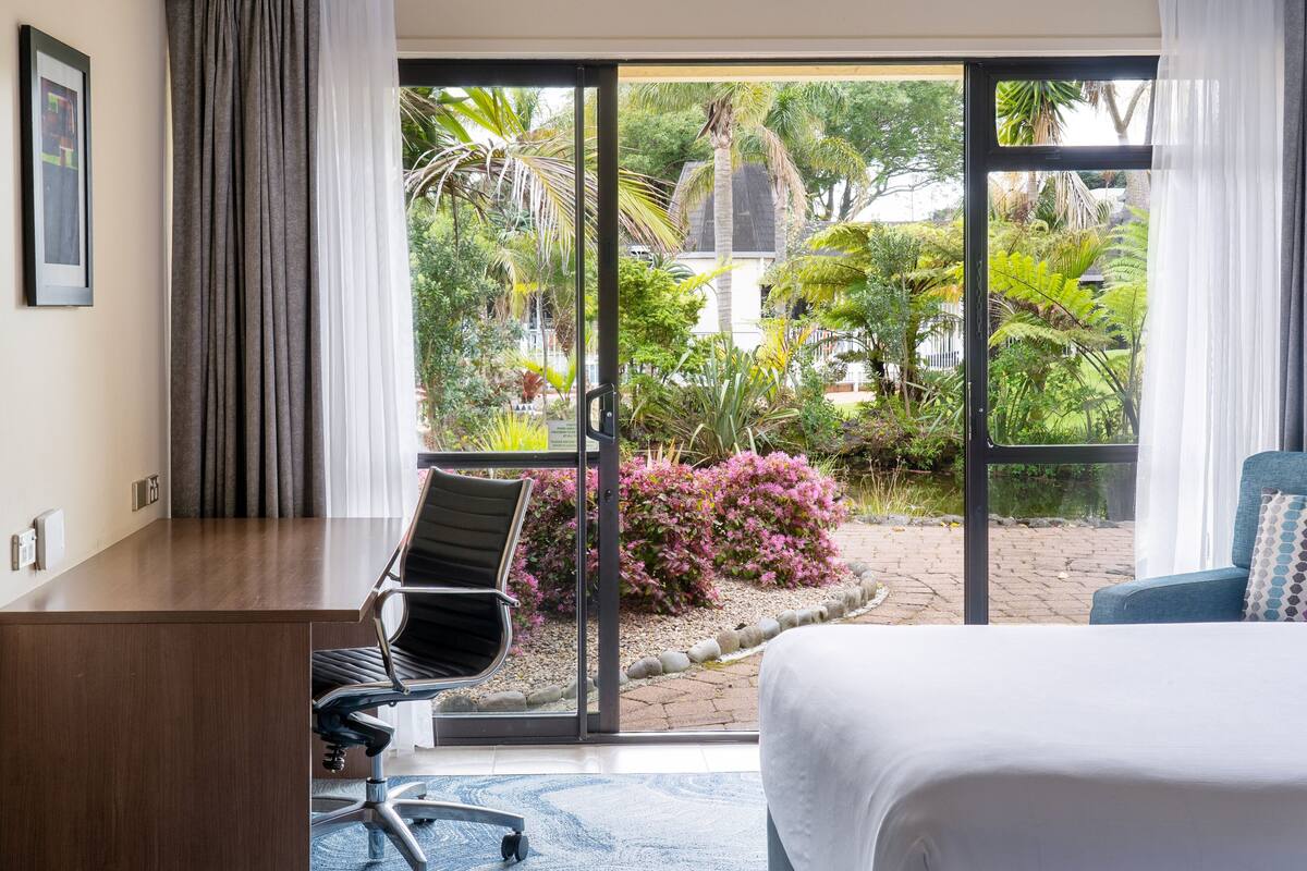 A hotel room with a bed, a desk, a chair, and a view of a garden through a glass door.
