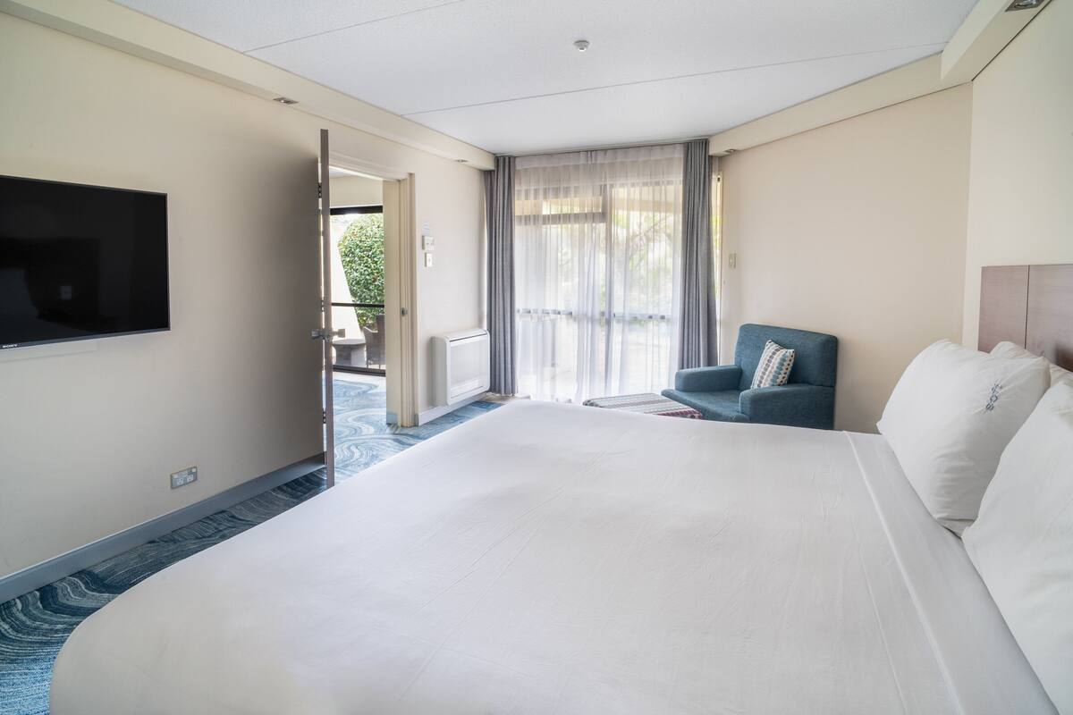 A hotel room with a large bed, a television mounted on the wall, a seating area with a chair, and a view through the door.