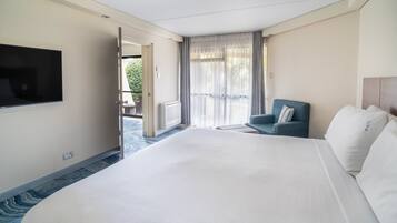 Habitación de hotel con una cama grande, televisión instalada en la pared, zona de estar con una silla y vistas a través de la puerta.