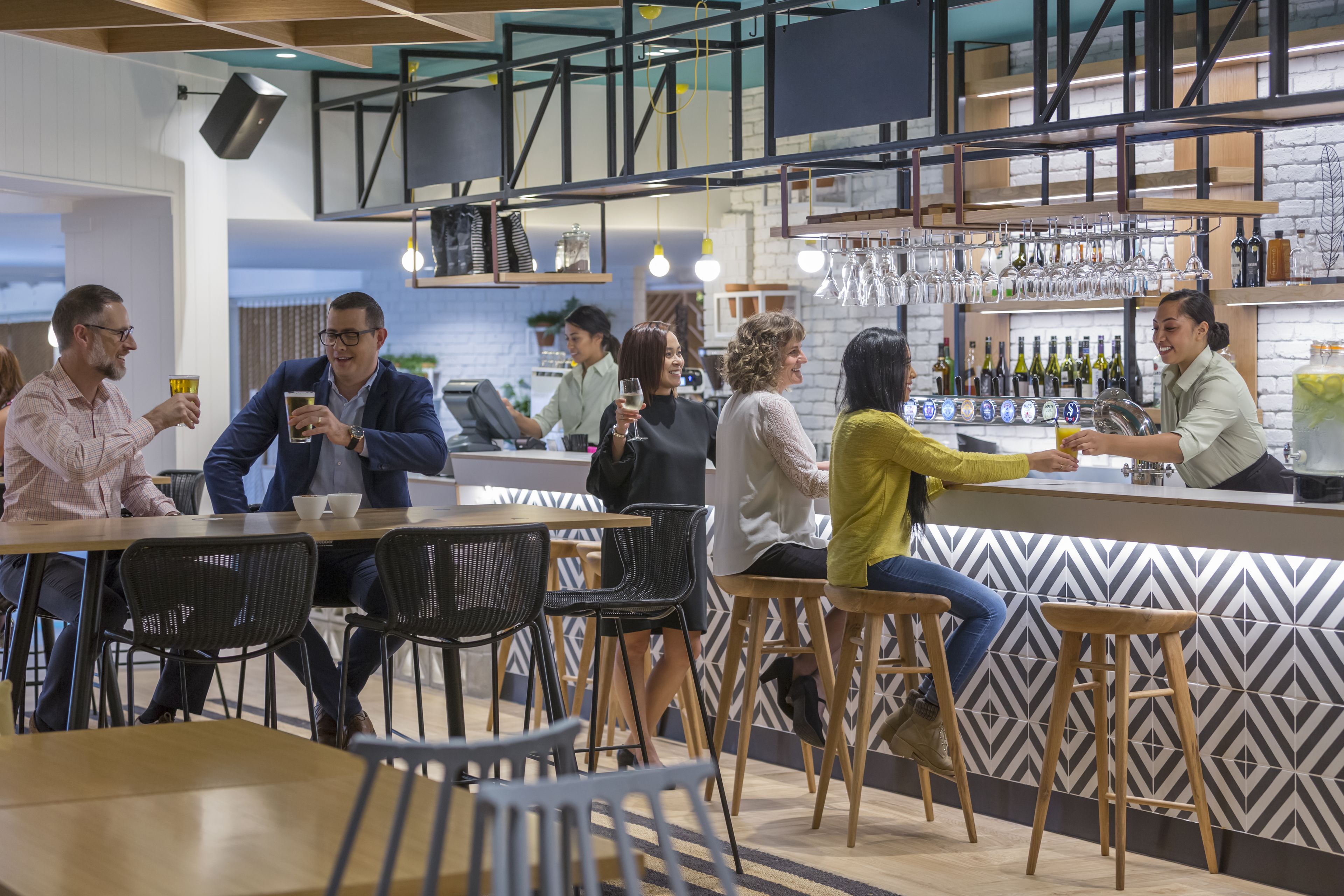A modern bar area with wooden stools, a bar counter, and a variety of bottles and glasses on shelves.
