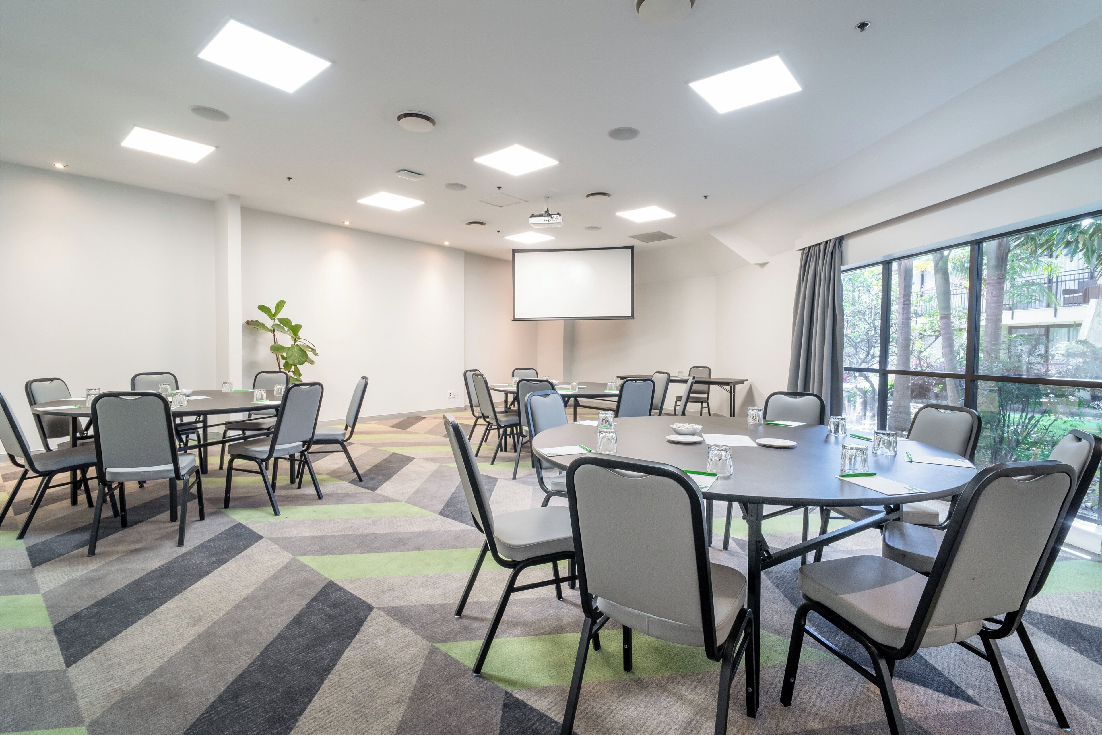 A conference room with multiple tables, chairs, a projector screen, and large windows.