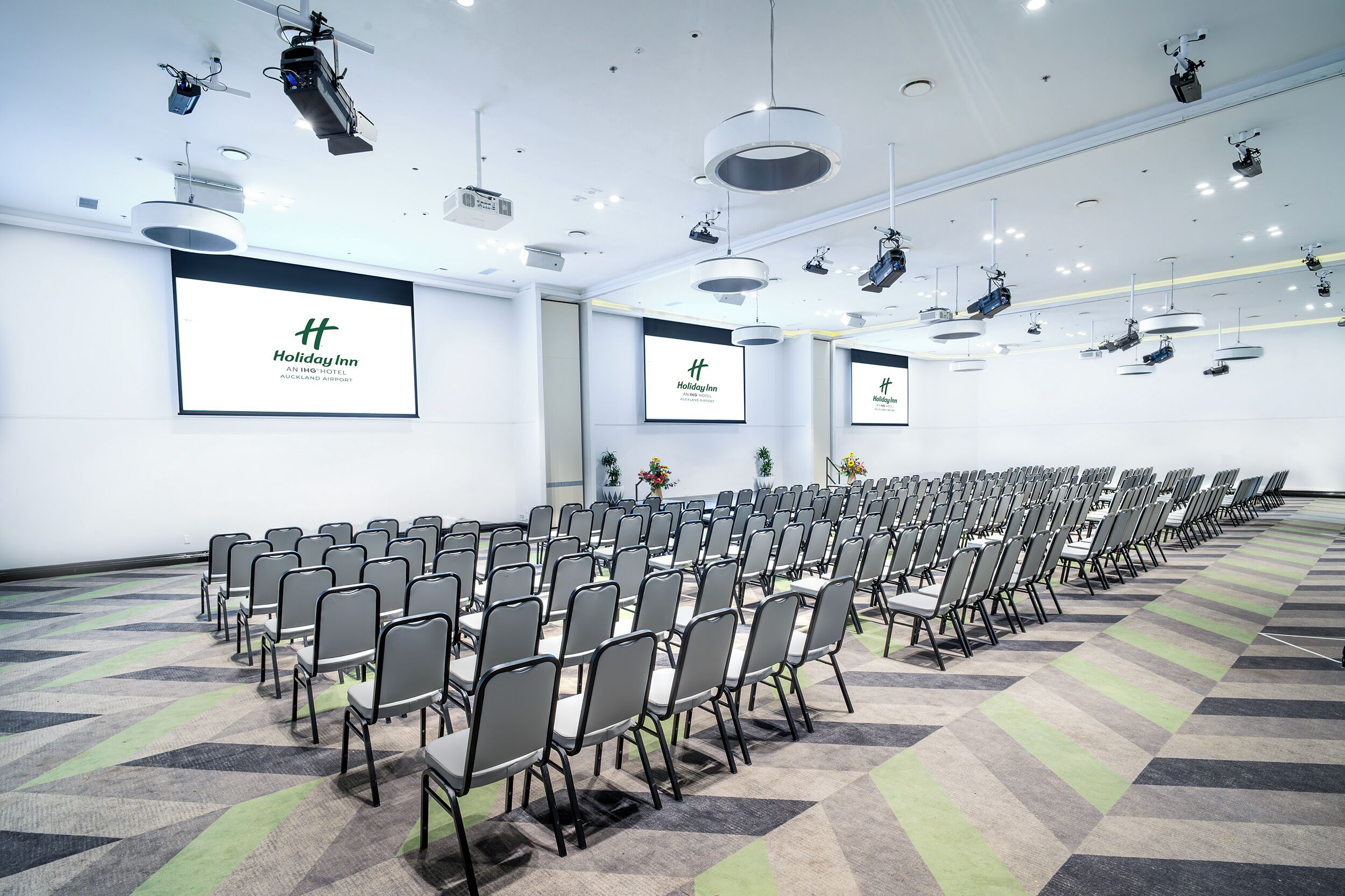 A large conference room with rows of chairs, a projector screen, and multiple ceiling-mounted lights.