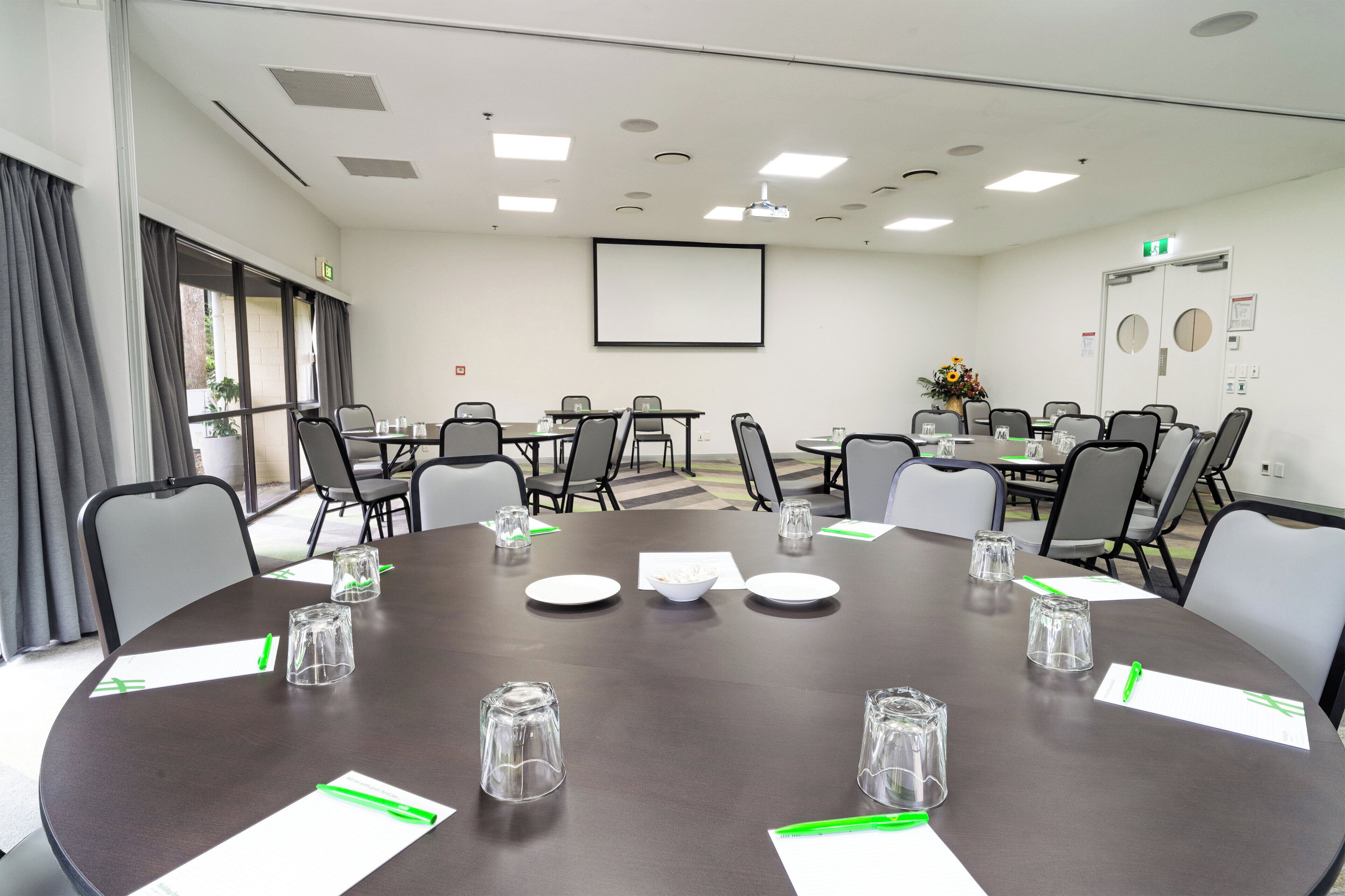 A conference room with a large table, chairs, a projector screen, and a window with curtains.