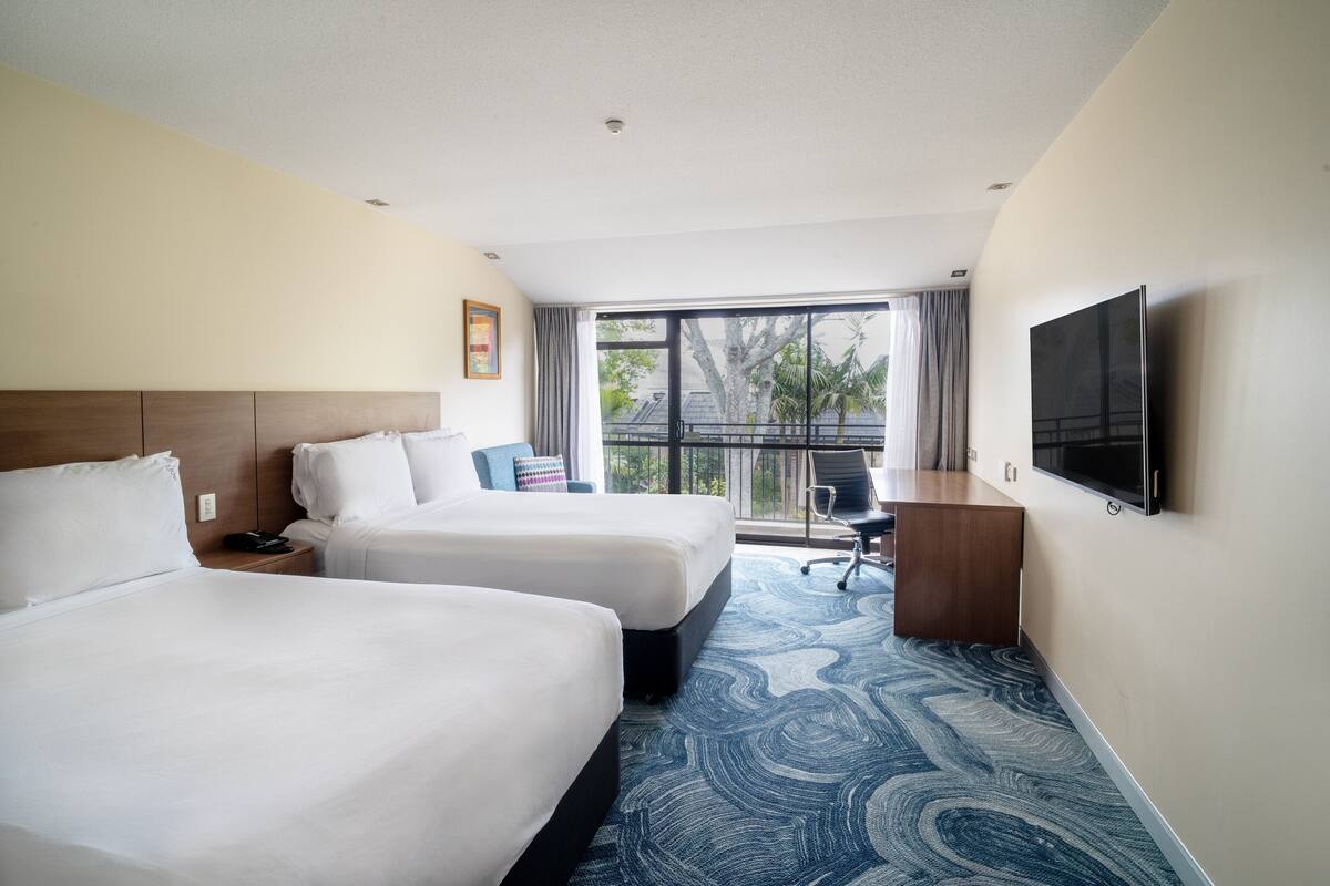 A hotel room with two beds, a desk, a chair, a TV, and a large window with a view.