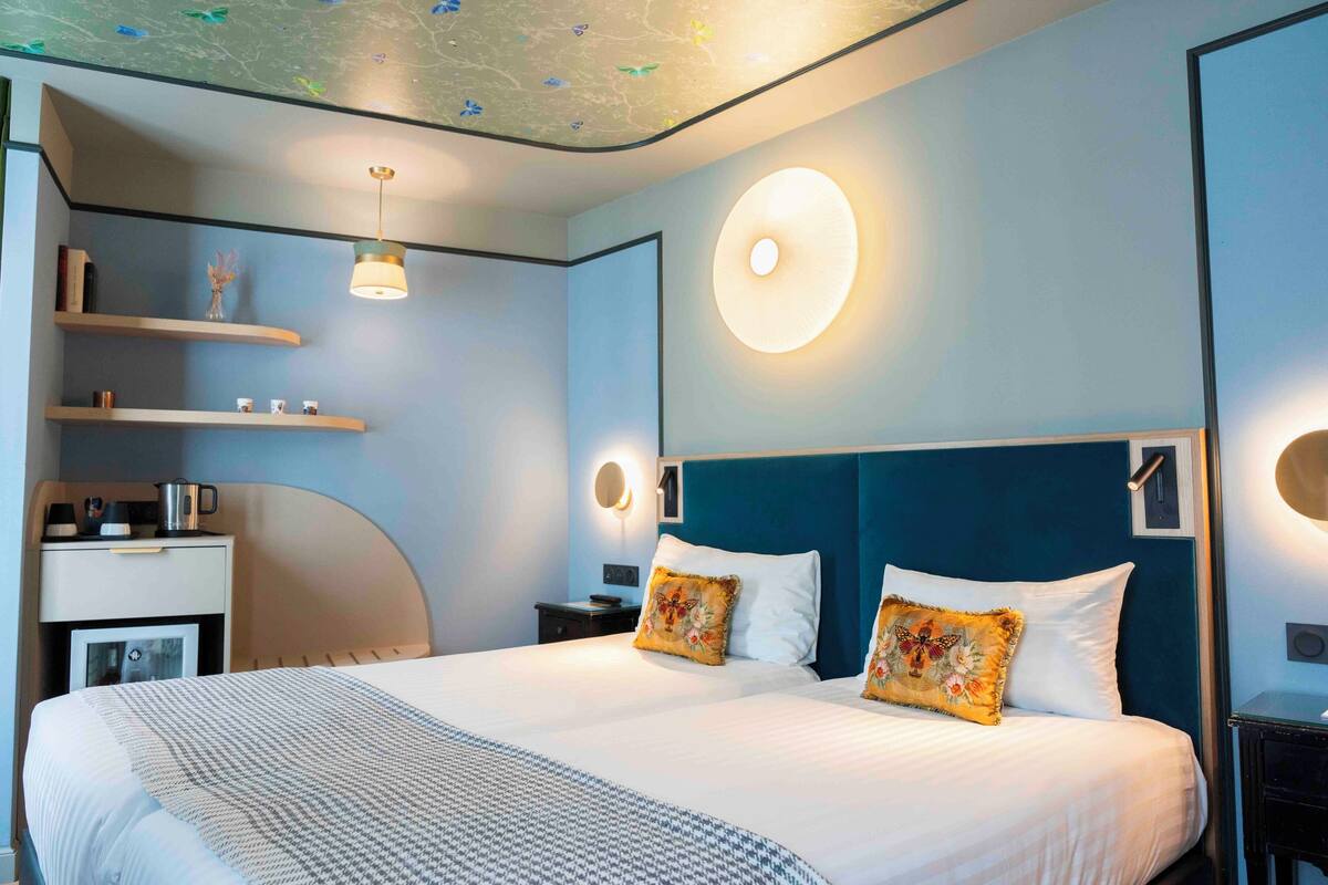 A modern hotel room with a large bed, a desk, and a chair. There are shelves with decorative items and a wall-mounted light fixture.