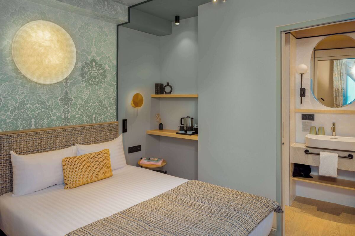 A bedroom with a large bed, a bedside table, a wall-mounted light, and a bathroom with a sink and mirror.