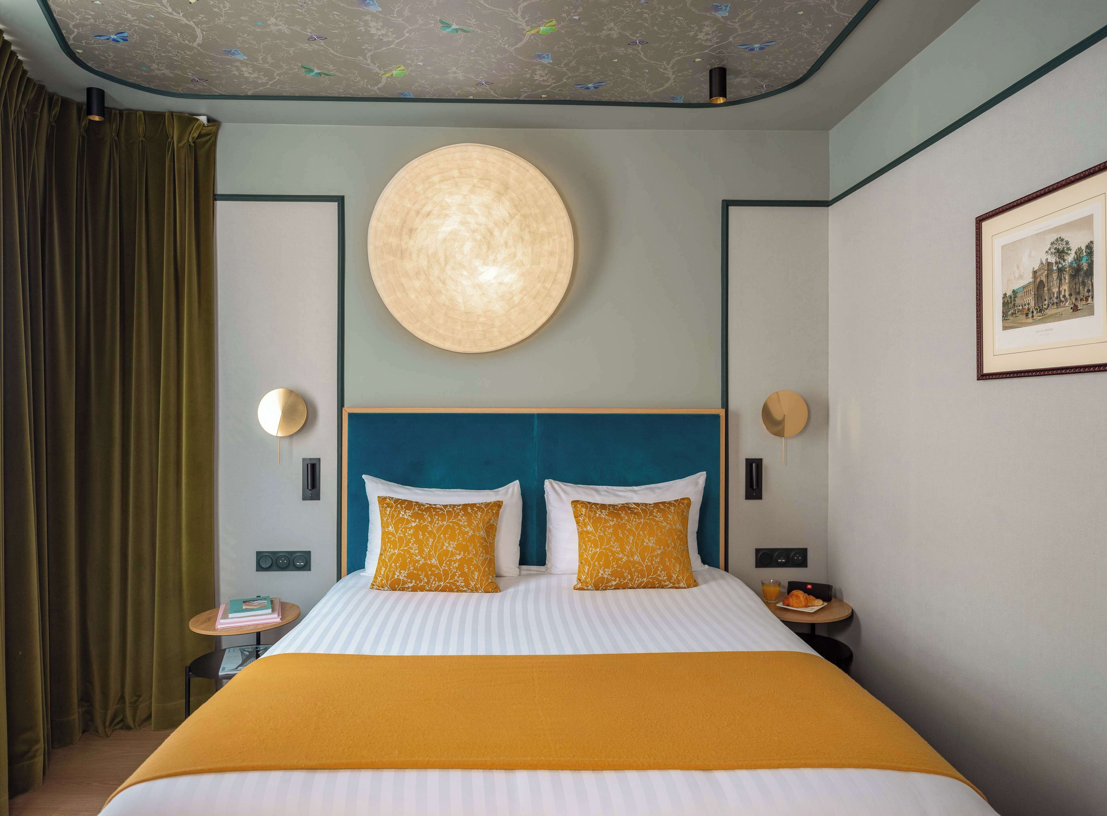 Habitación de hotel con una cama grande, dos mesitas de noche, una lámpara de pared redonda y un cuadro enmarcado.
