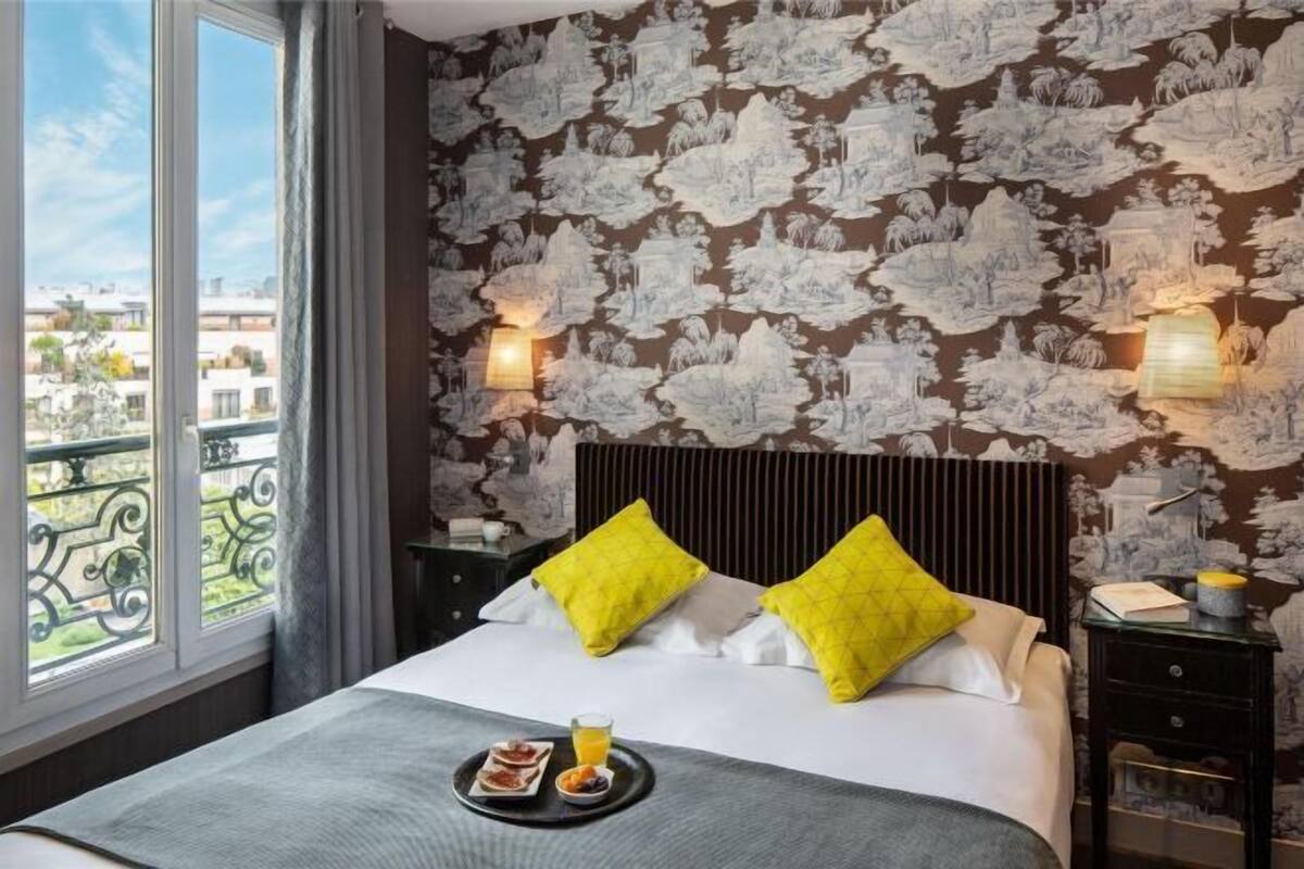 A hotel room with a large window, a bed with yellow pillows, a bedside table with a lamp, and a tray with food and a glass of orange juice.