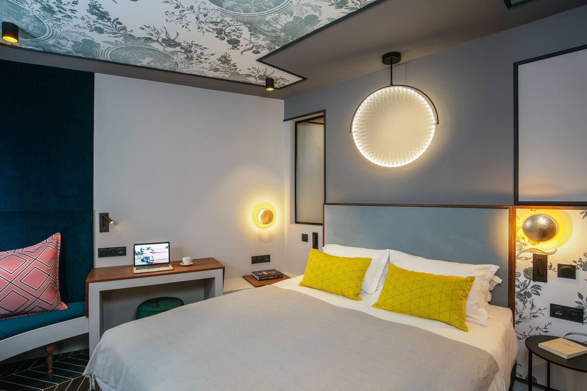 A modern hotel room with a large bed, a desk with a laptop, and a round wall light.
