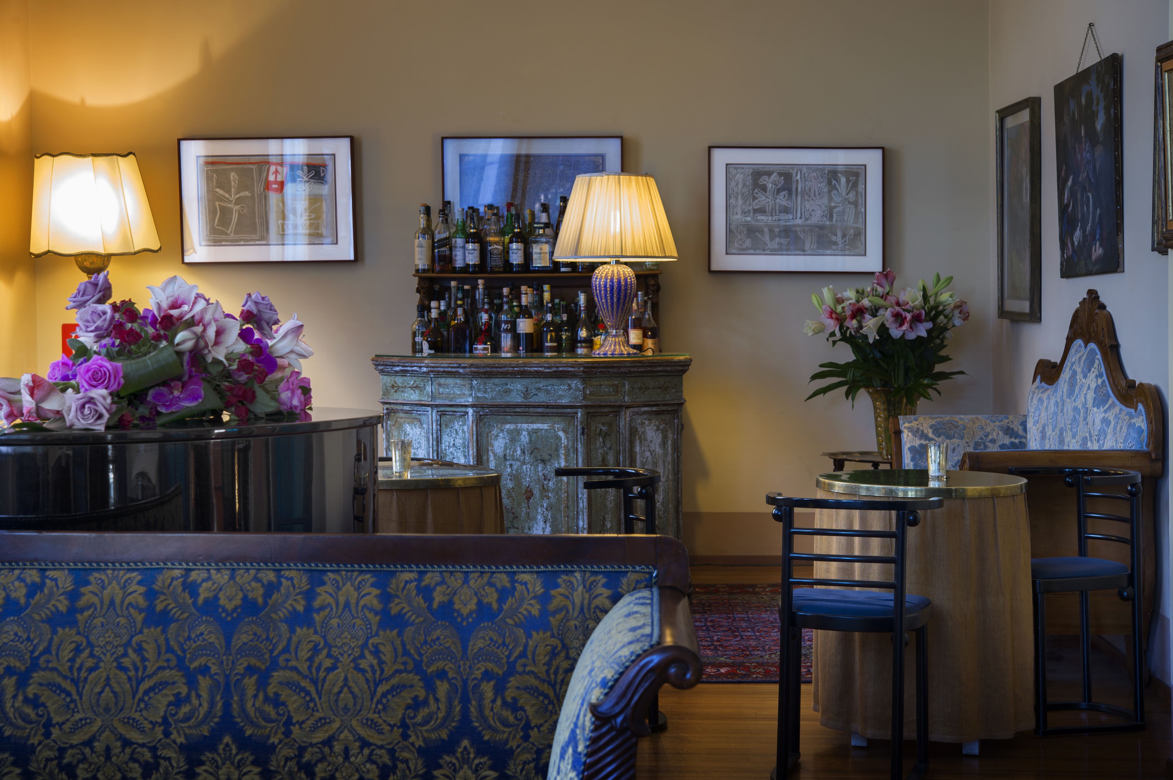 A room with a bar area, a sofa, and two chairs. There are framed pictures on the wall and a lamp on the bar.