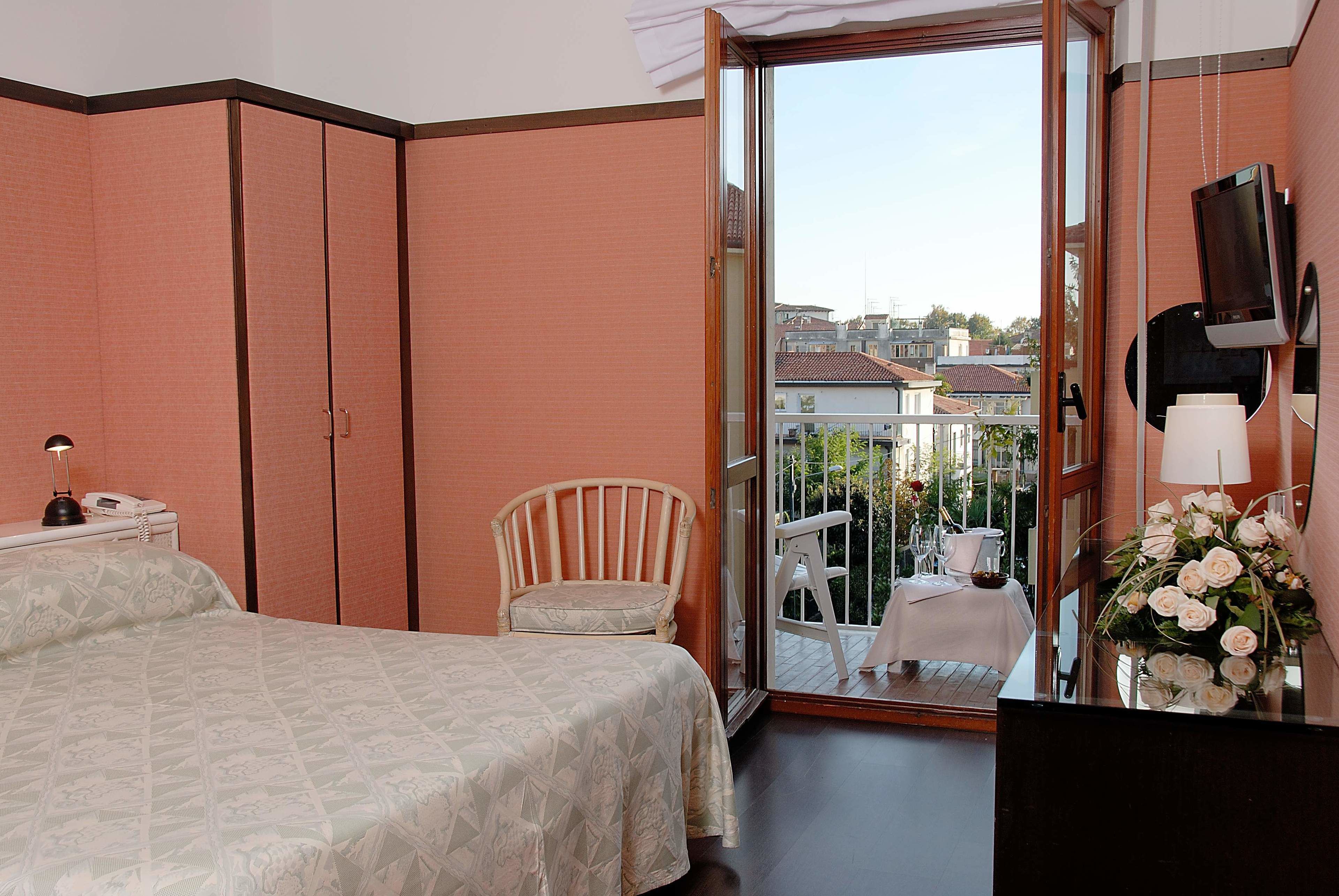 A bedroom with a bed, a chair, a desk with a lamp, a TV, and a balcony with a table and chairs.