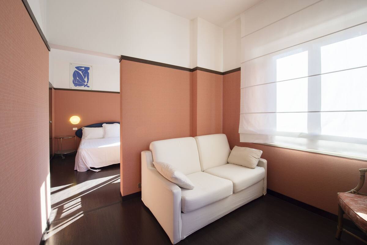 A hotel room with a white sofa, a bed with white bedding, a small table, and a window with blinds.