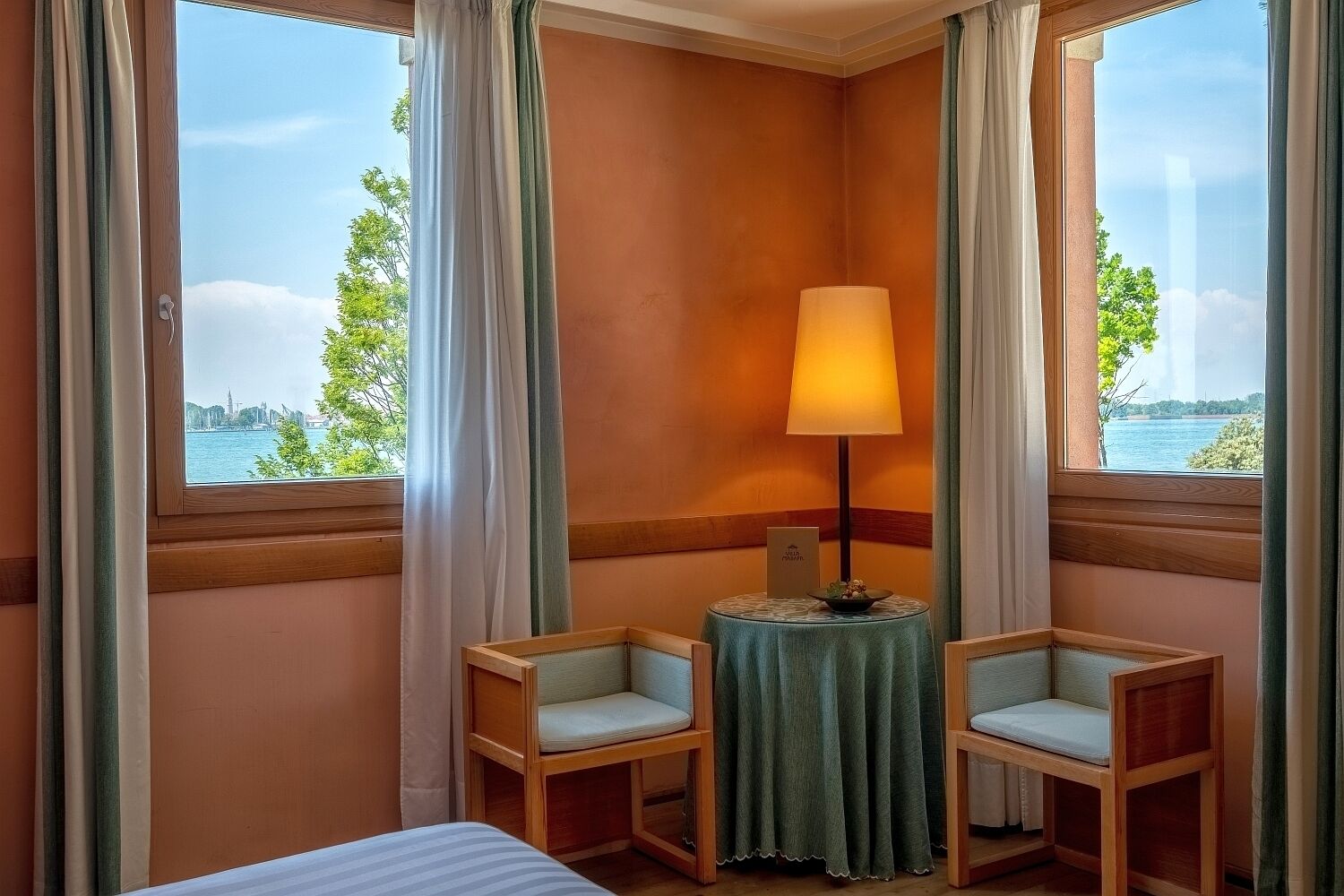 A hotel room with a bed, two armchairs, a small table with a lamp, and a view of a body of water and trees.