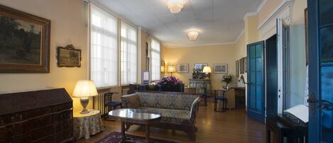 A spacious living room with a sofa, a coffee table, and a patterned rug. There is a fireplace with a mantel, a large painting on the wall, and a chandelier.