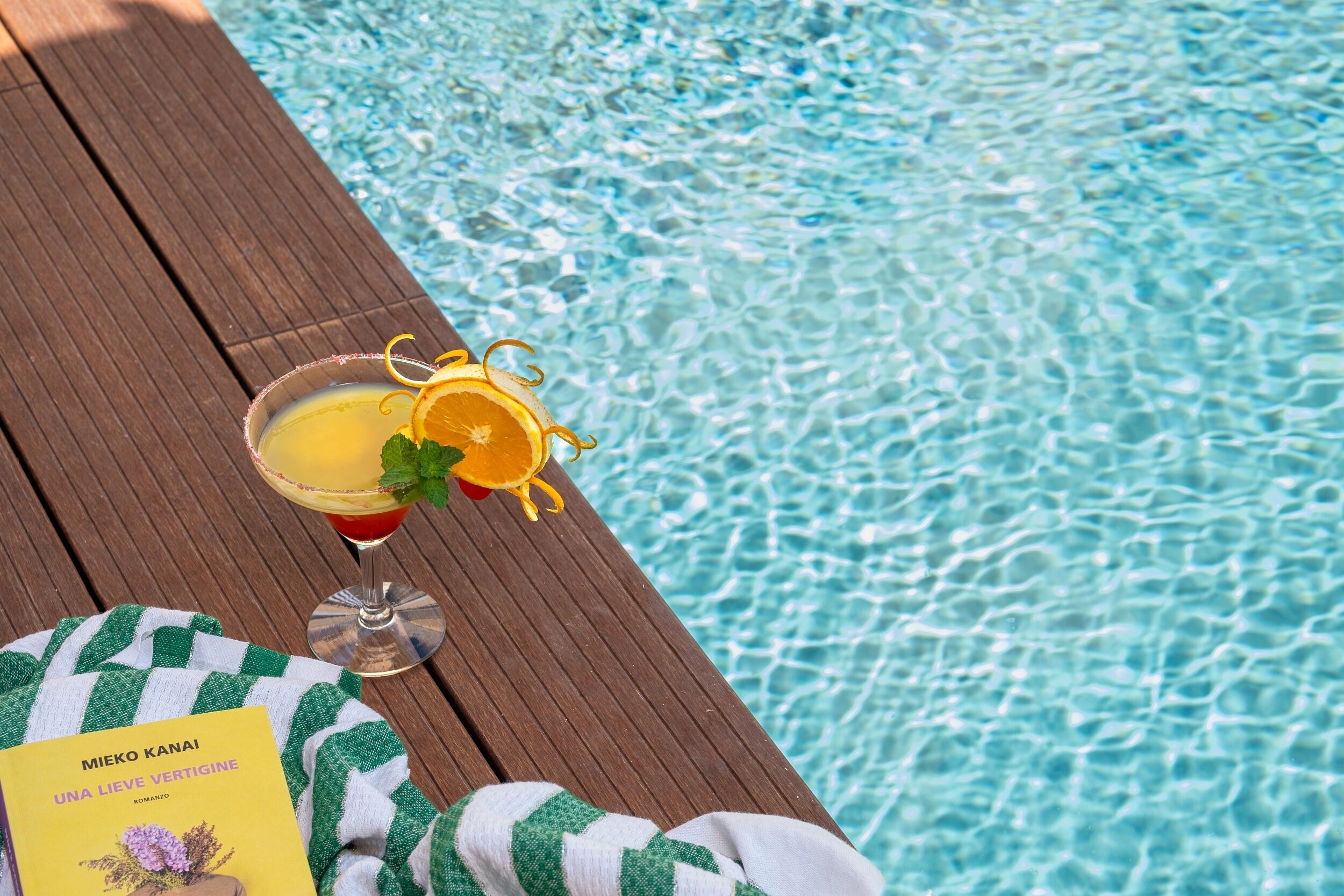 Un'area prendisole in legno accanto a una piscina, con un drink, un libro e un asciugamano.