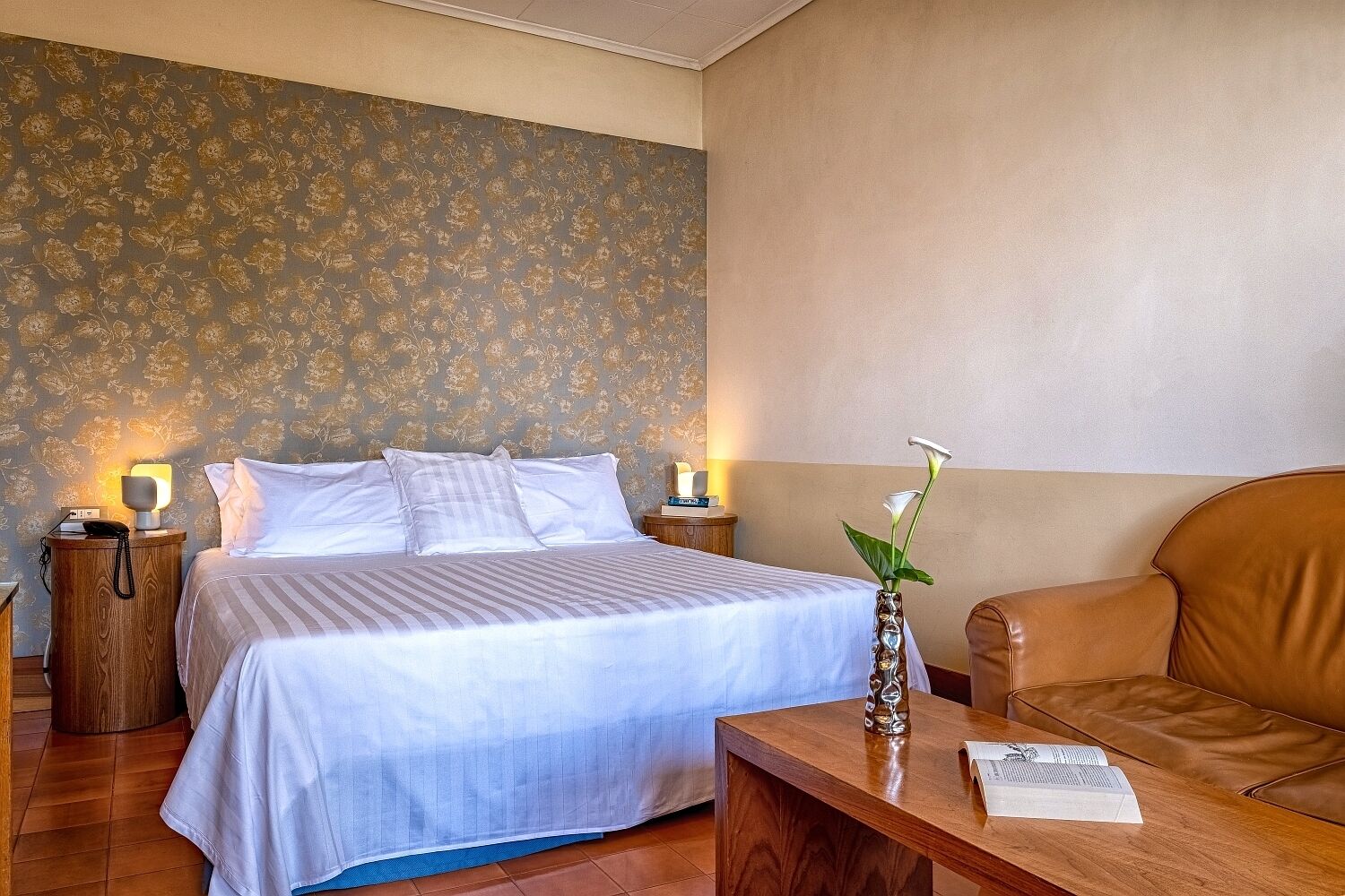 Ein Hotelzimmer mit einem Bett, Nachttischen, einem kleinen Tisch, einem Holztisch, einem Ledersessel und einer Vase mit Blumen.
