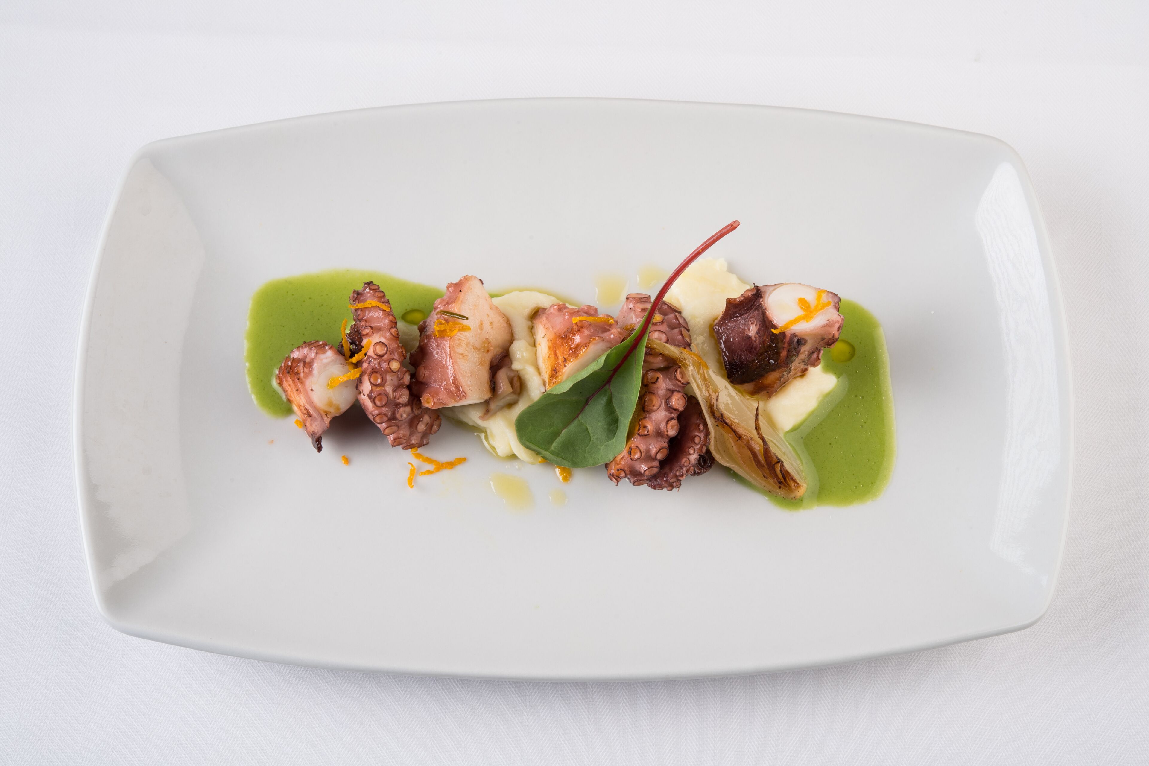A white rectangular plate with octopus pieces, green sauce, and a garnish.