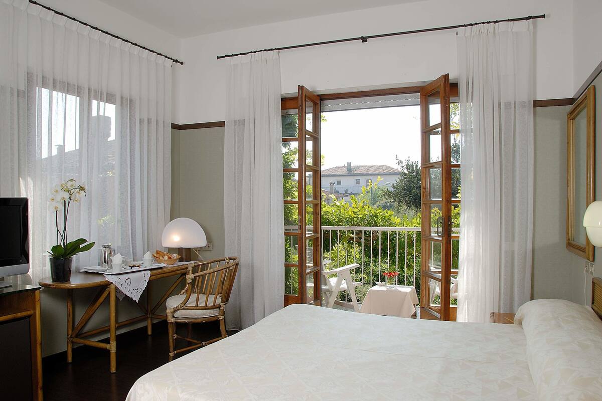 A hotel room with a bed, a desk with a lamp, a chair, and a balcony with a table and chairs.