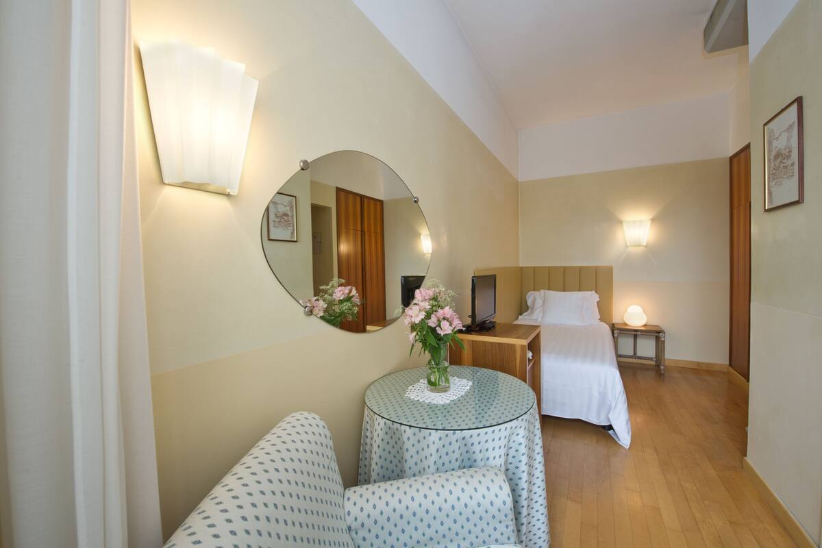 A hotel room with a bed, bedside table, lamp, mirror, and a small round table with a vase of flowers.