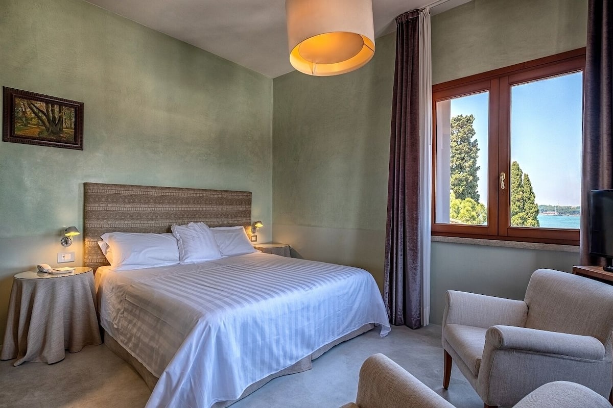 A hotel room with a large bed, a view of the sea, a television, and a chair.
