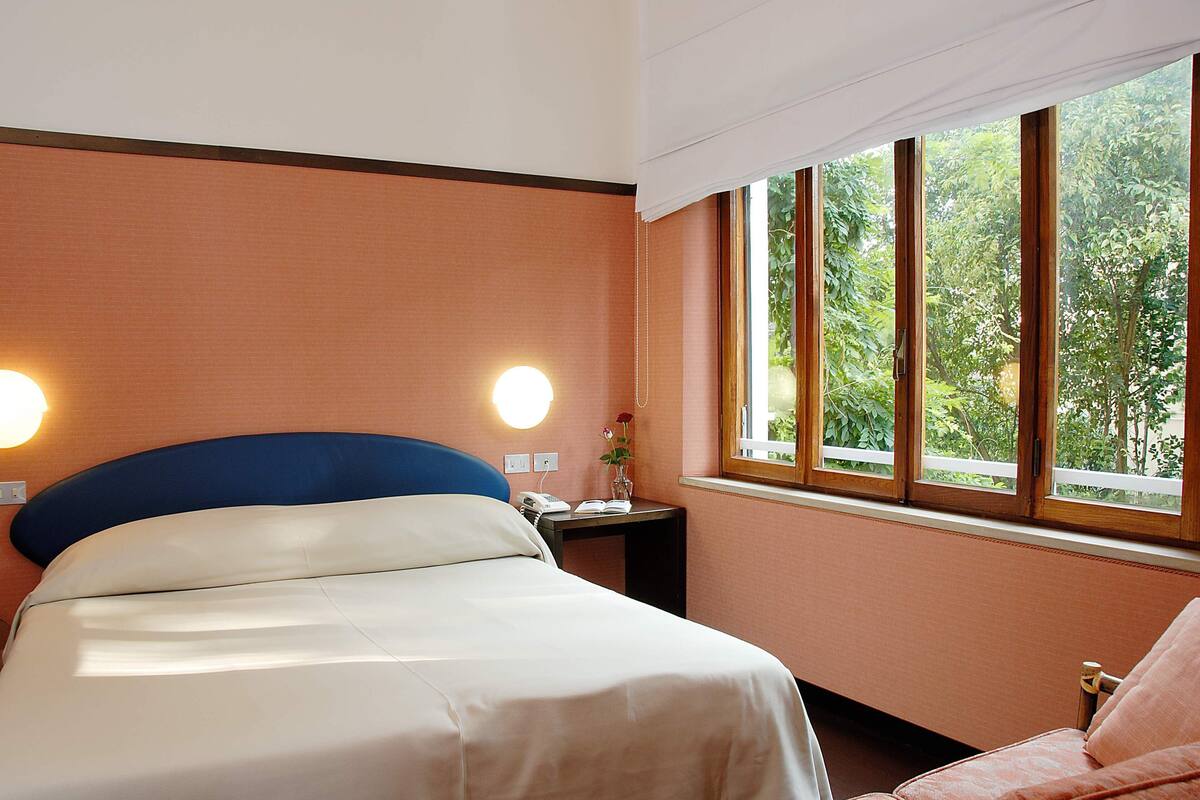 A hotel room with a bed, bedside table, lamp, and a window with a view of greenery.