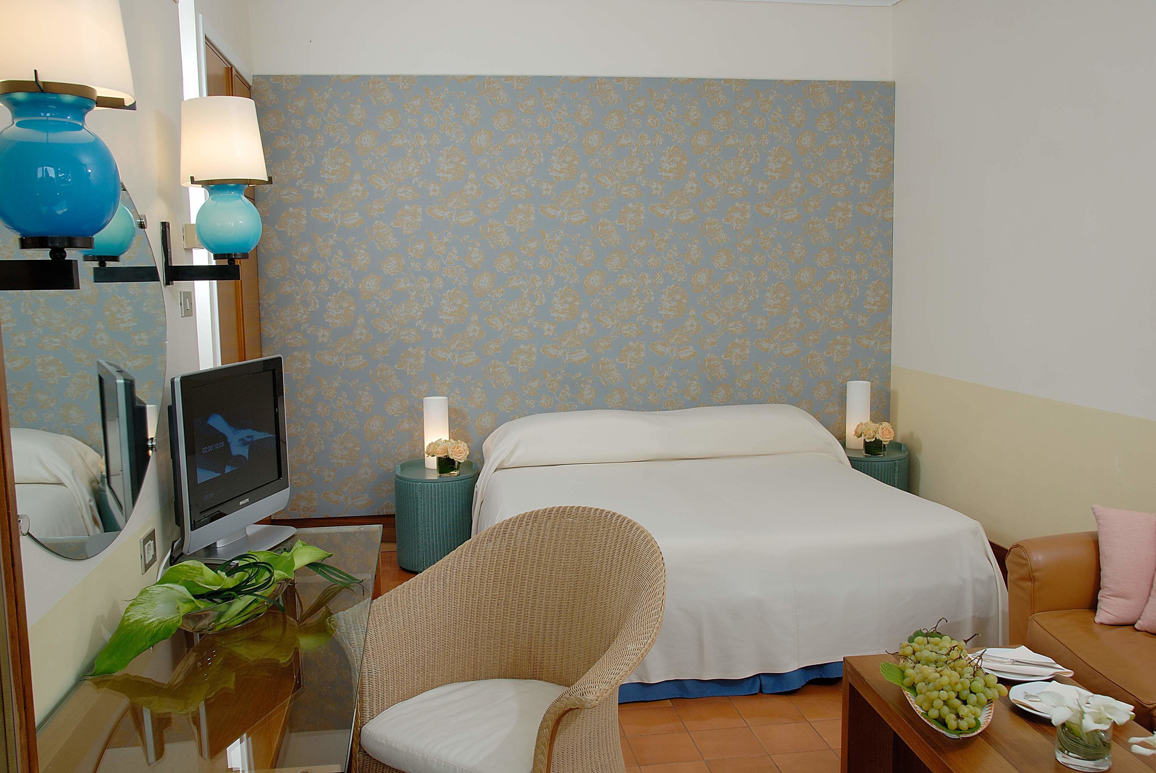 Una habitación de hotel con una cama, una silla, un televisor, una mesita de noche con una lámpara y una pequeña mesa con un frutero.