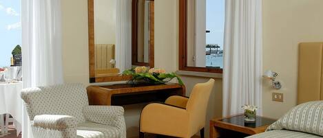 A hotel room with a bed, a desk, a chair, a mirror, and a window with a view of the sea.