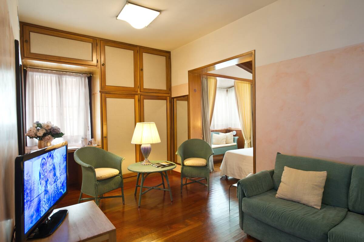 A hotel room with a green sofa, two armchairs, a small table with a lamp, a TV, and a bed with a white duvet.