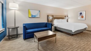 Une chambre d’hôtel moderne avec un canapé bleu, une table basse en bois, un lit avec du linge de lit blanc, une table de chevet, une lampe et des œuvres d’art abstraites accrochées au mur.