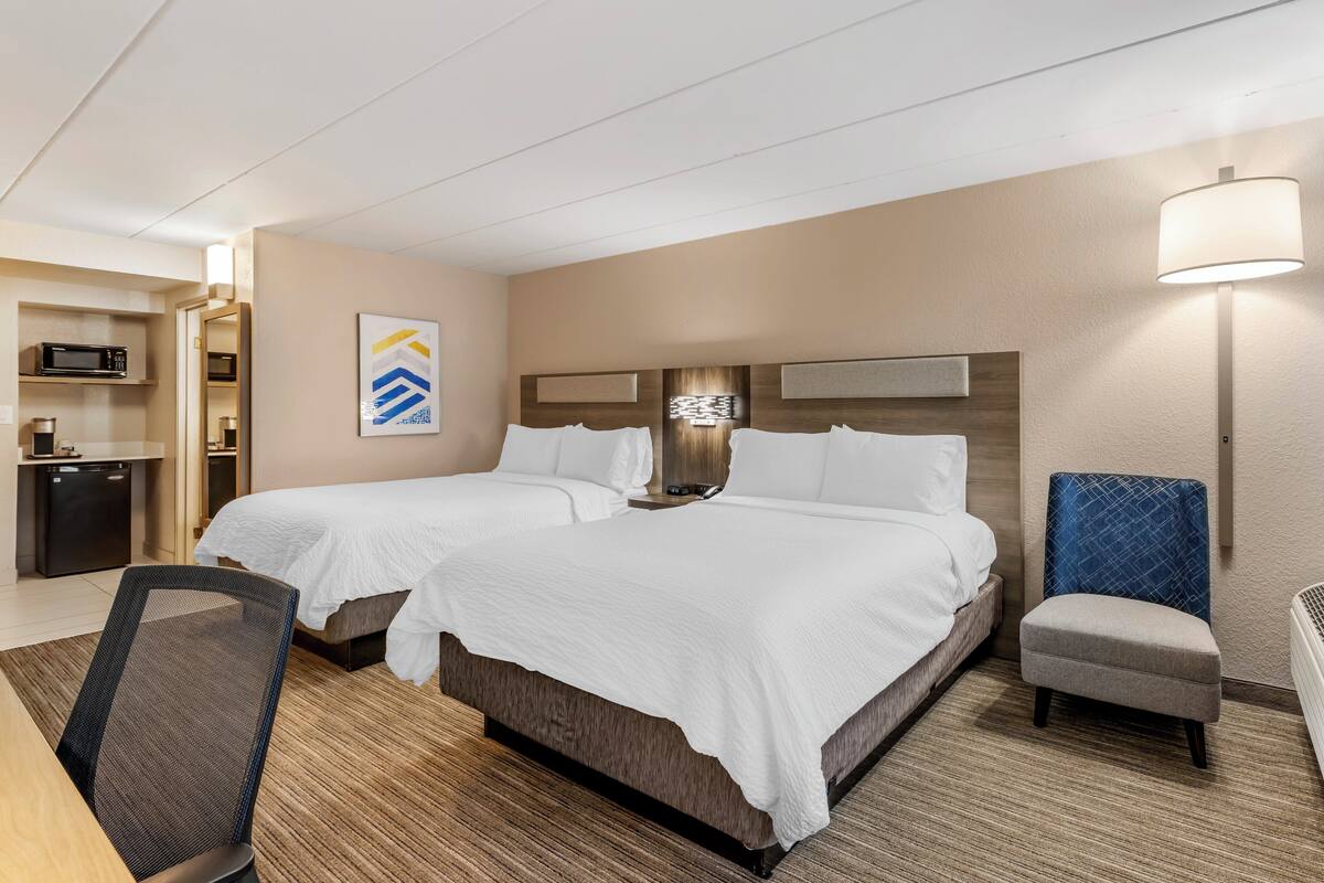 A hotel room with two beds, a desk, a chair, a microwave, and a lamp.