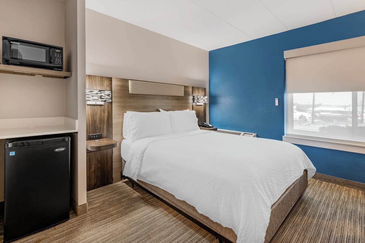 A hotel room with a bed, a microwave, a mini fridge, a wooden headboard, a window with a view, and a blue accent wall.