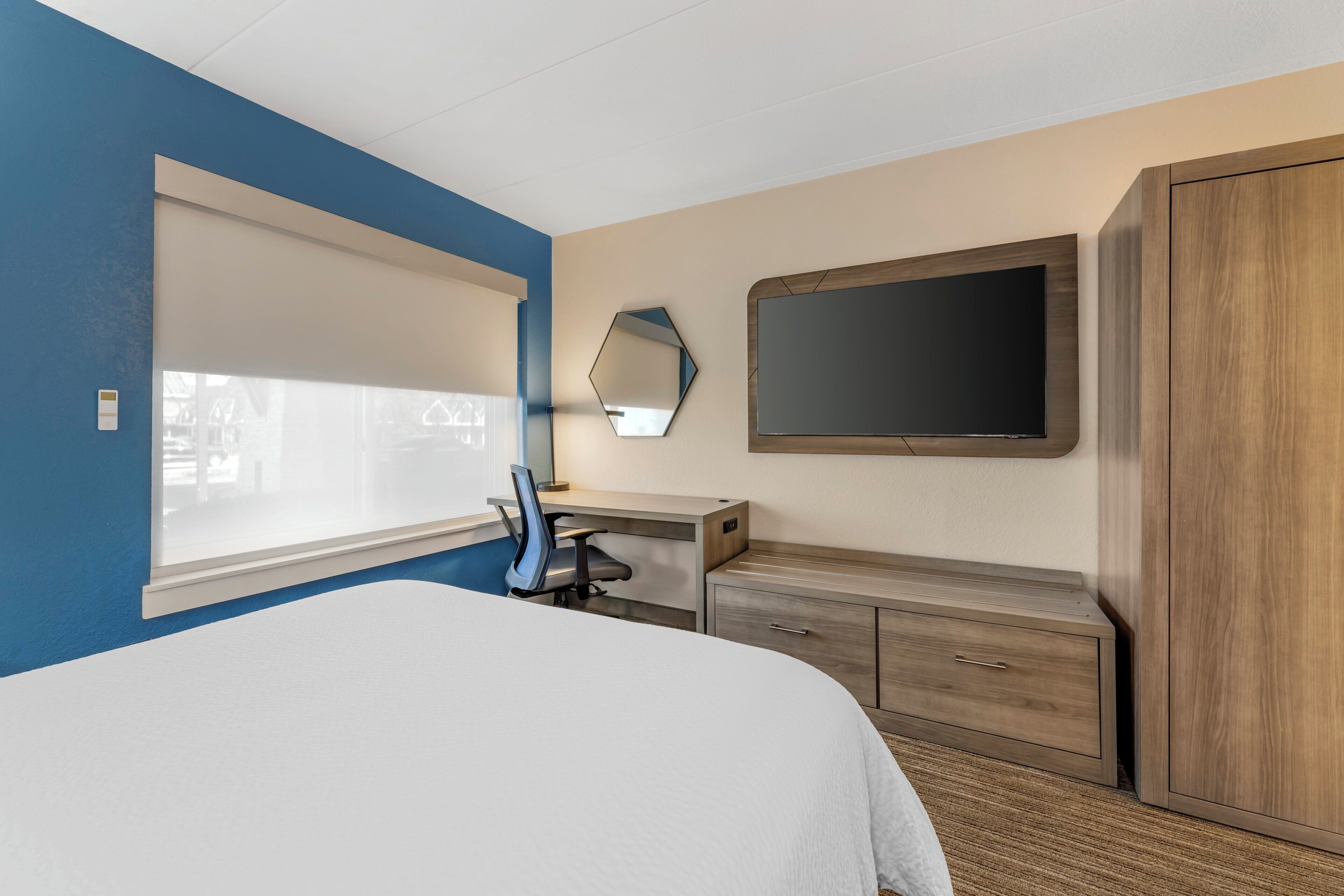 A hotel room with a bed, a desk, a chair, a wardrobe, a mirror, and a TV.
