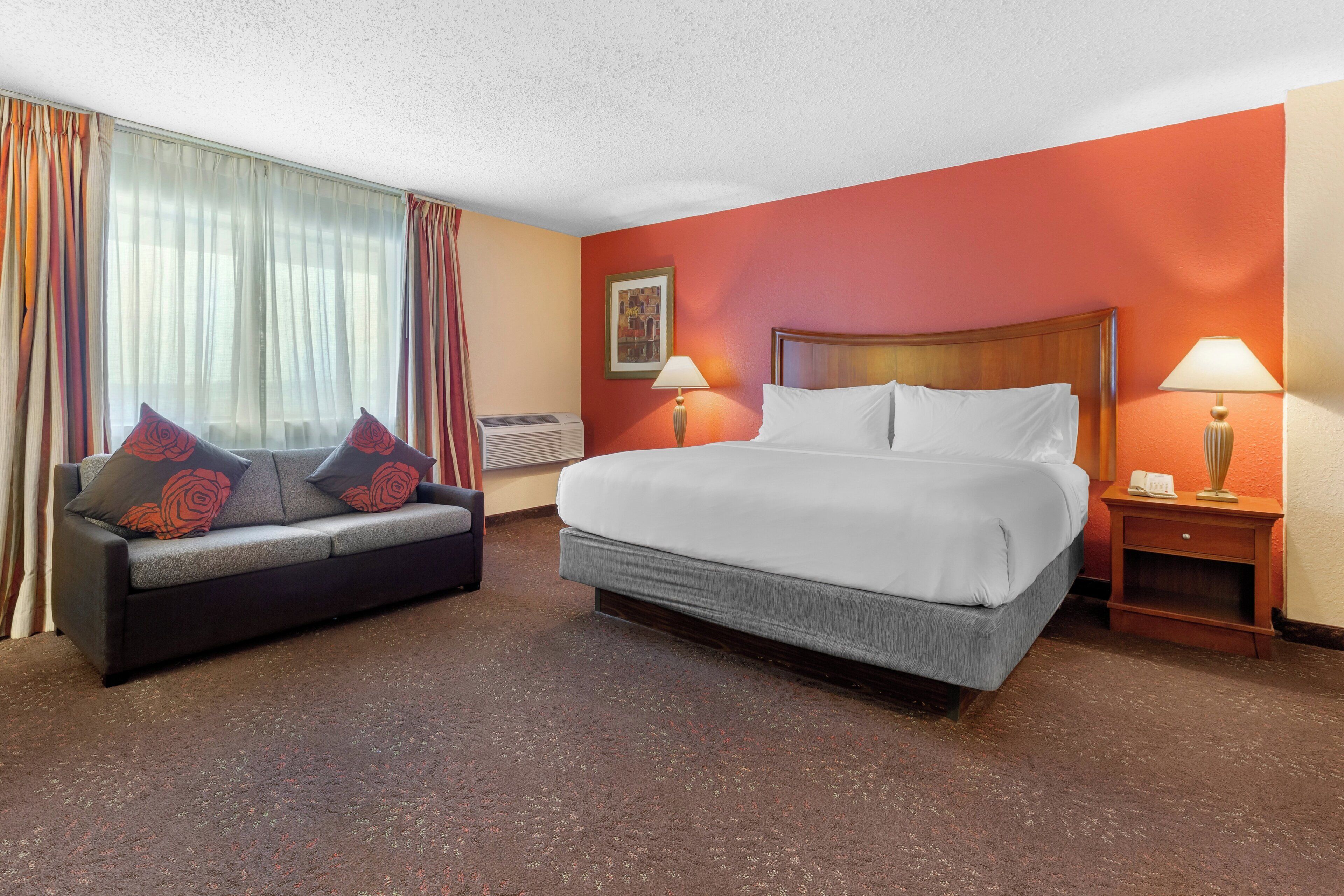 A hotel room with a bed, a sofa, two bedside tables, a lamp, and a window with curtains.