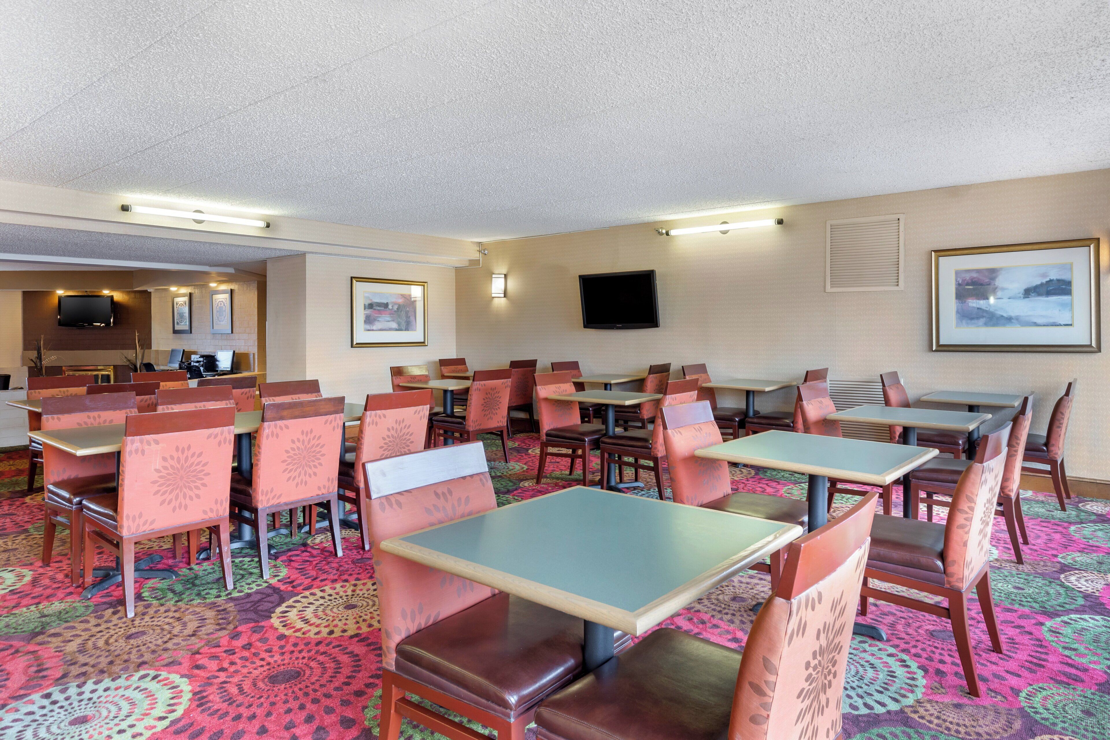 A hotel dining area with multiple tables and chairs, a flat-screen TV mounted on the wall, and framed artwork on the walls.