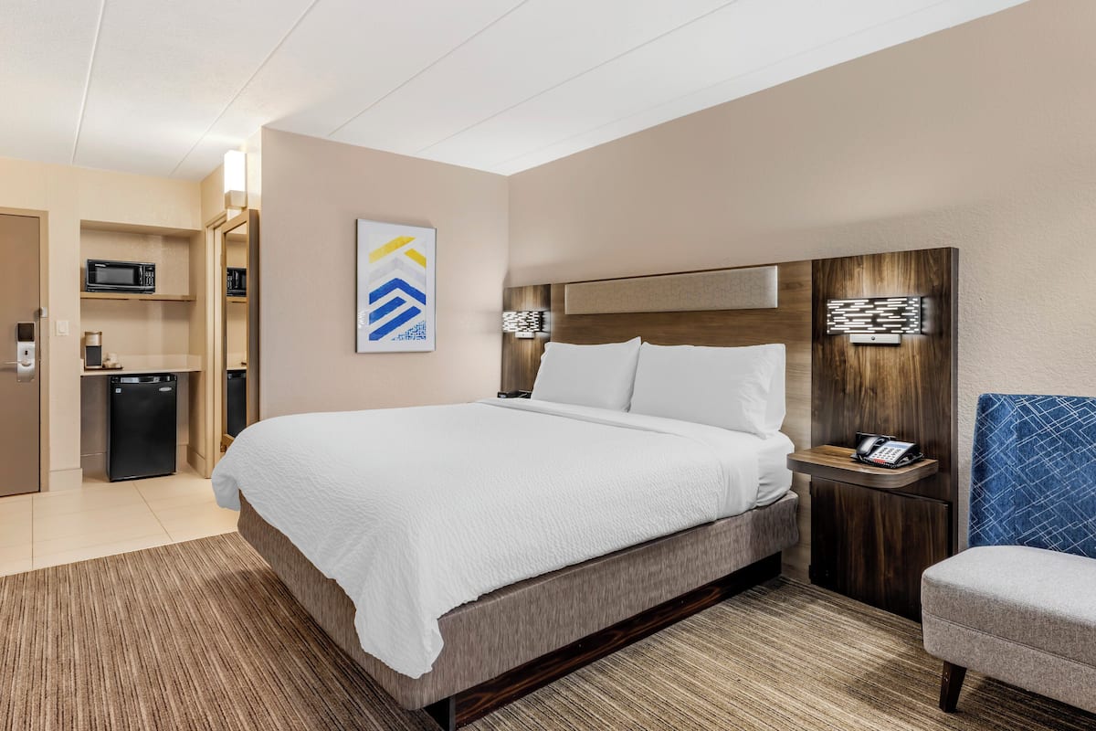 A hotel room with a bed, bedside table, chair, microwave, refrigerator, and a wall-mounted artwork.