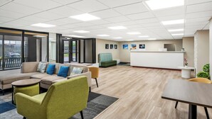 A spacious lounge area with a sectional sofa, a round table, and a green armchair. There is a reception desk in the background with a small plant on it. - Red Roof Inn Perryville (Perryville)