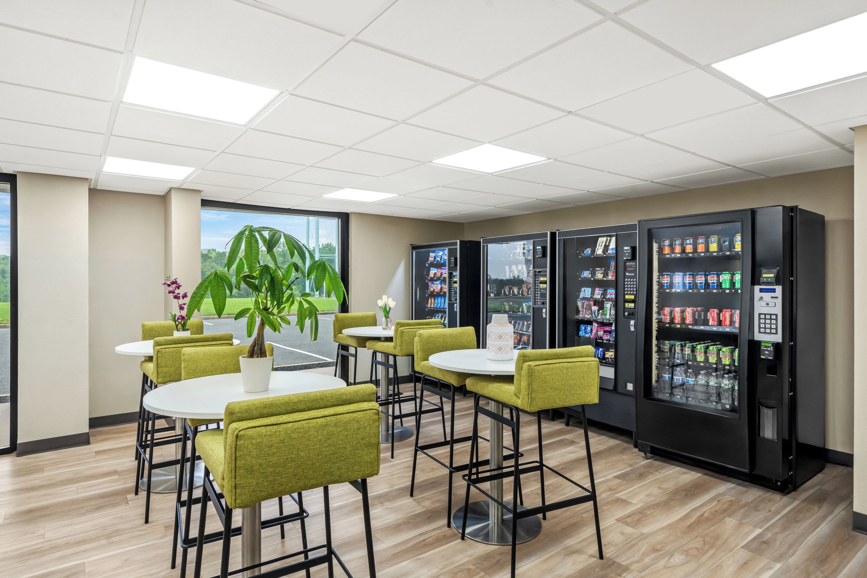 A modern indoor space with round tables and green chairs, vending machines, and a large potted plant.