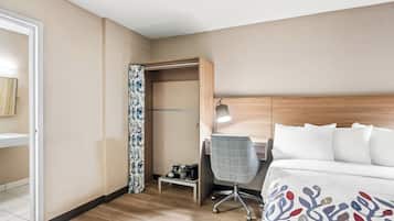 Habitación de hotel con cama, escritorio y silla. Se ve un armario con espejo y una mesita de noche con lámpara.