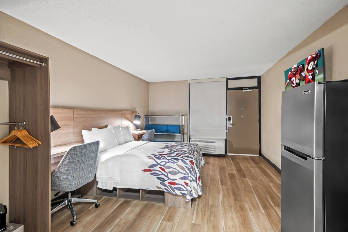 A hotel room with a bed, desk, chair, and a refrigerator.
