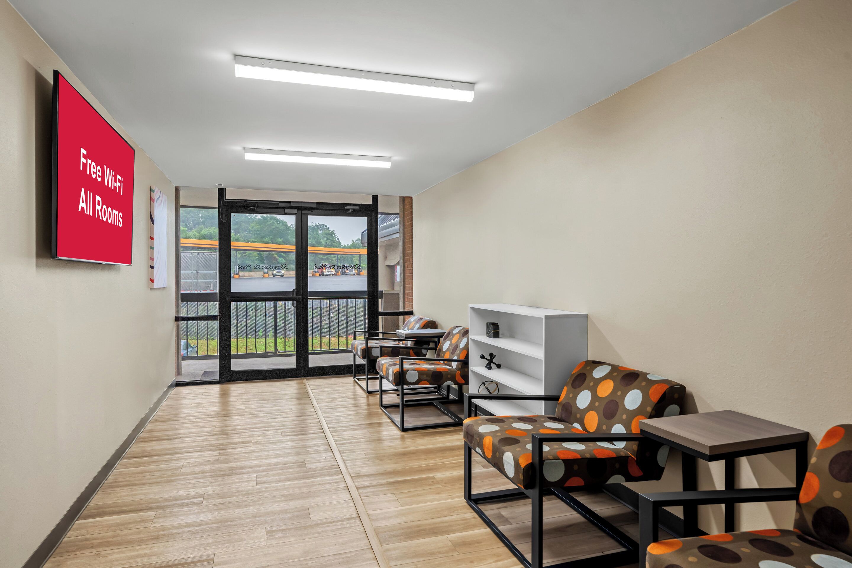 A modern hotel room with a wooden floor, a large window with a view of the outdoors, a flat-screen TV on the wall, and a seating area with a white console table and chairs.