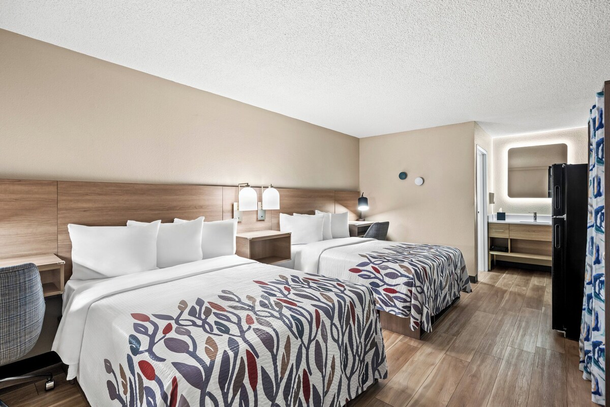 A hotel room with two beds, a desk, a chair, a desk lamp, a wall-mounted TV, and a refrigerator.