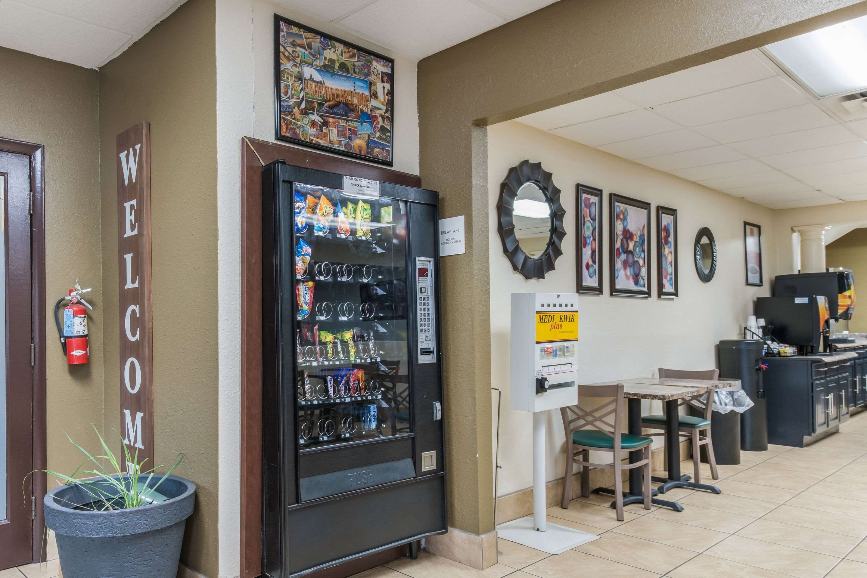 A hotel lobby with a vending machine, a fire extinguisher, a welcome sign, a breakfast sign, and a mirror on the wall.