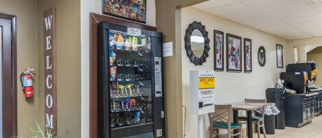 A hotel lobby with a vending machine, a fire extinguisher, a welcome sign, a breakfast sign, and a mirror on the wall.